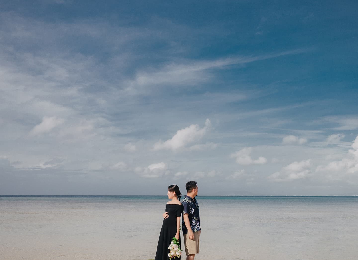 Expecting couple stand in shallow water under a vast Okinawa sky