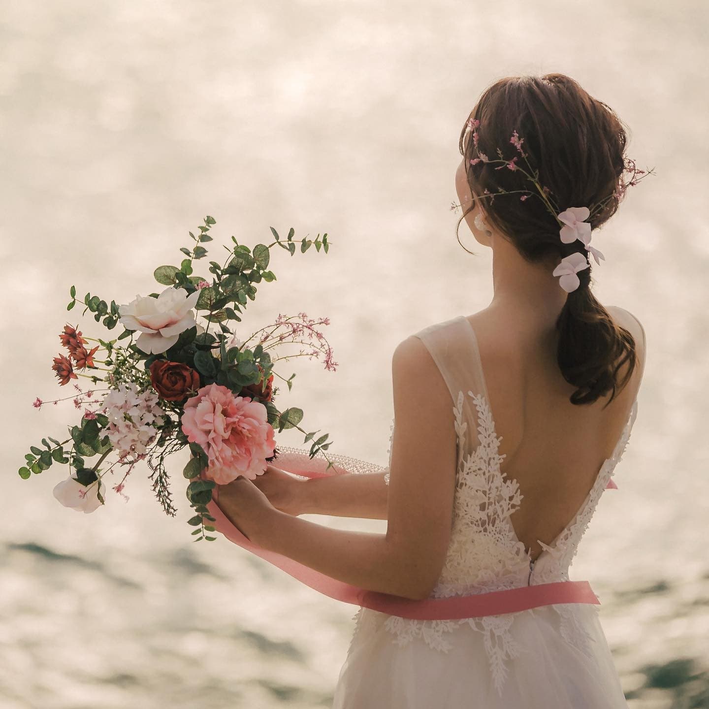 Back view of a bride holding a pink bouquet before a softly shimmering sea