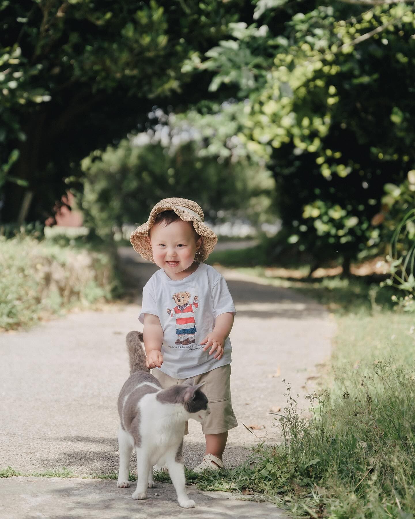 Smiling toddler in a straw hat with a cat on a shady path in Okinawa