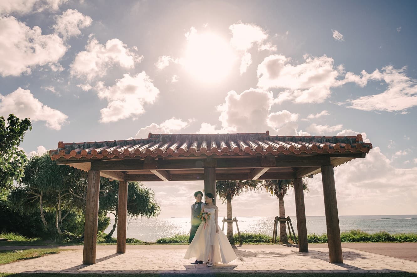 Couple beneath a red-tiled Okinawan pavilion with the sun blazing above the sea