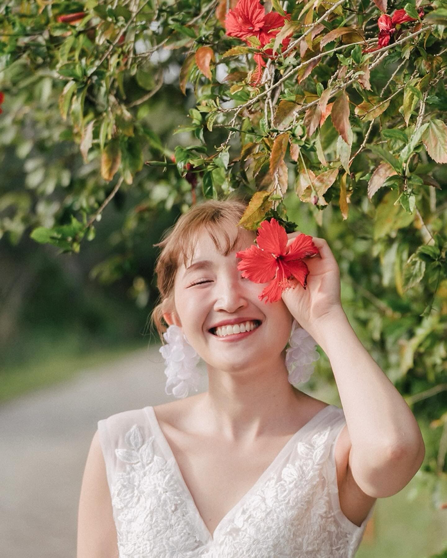 Smiling bride holds a red hibiscus to her eye amid Okinawa greenery