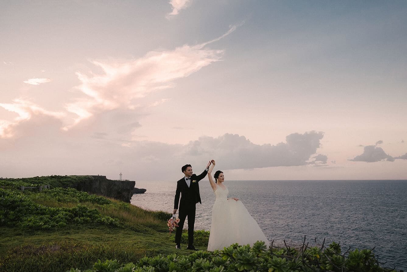 桃色の夕焼け雲の岬で手を取り合う新郎新婦、海を望む沖縄ロケーション
