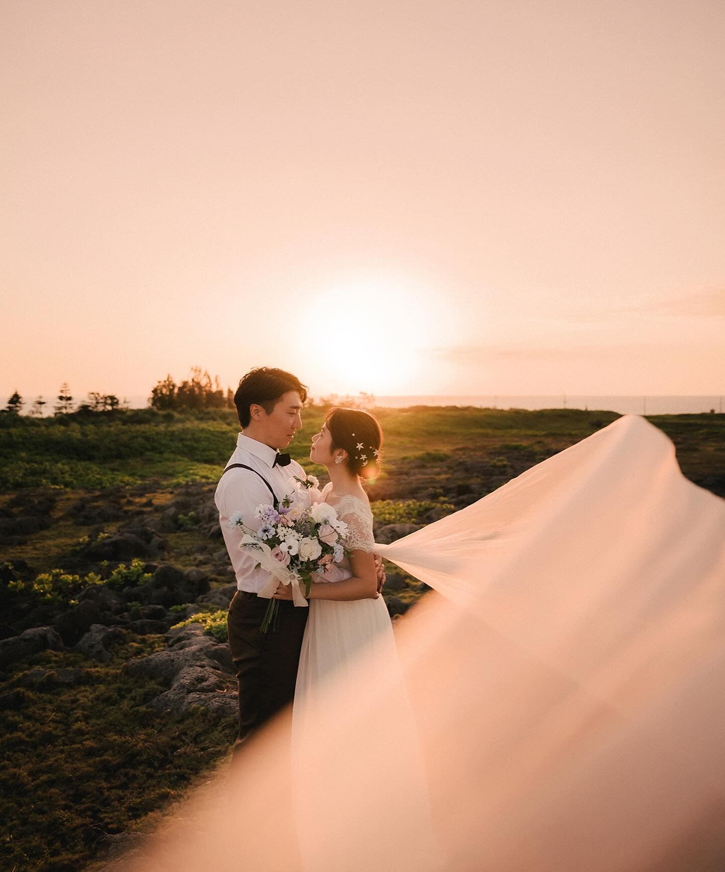 Couple face to face on an Okinawa hillside, the veil sweeping in the sunset light