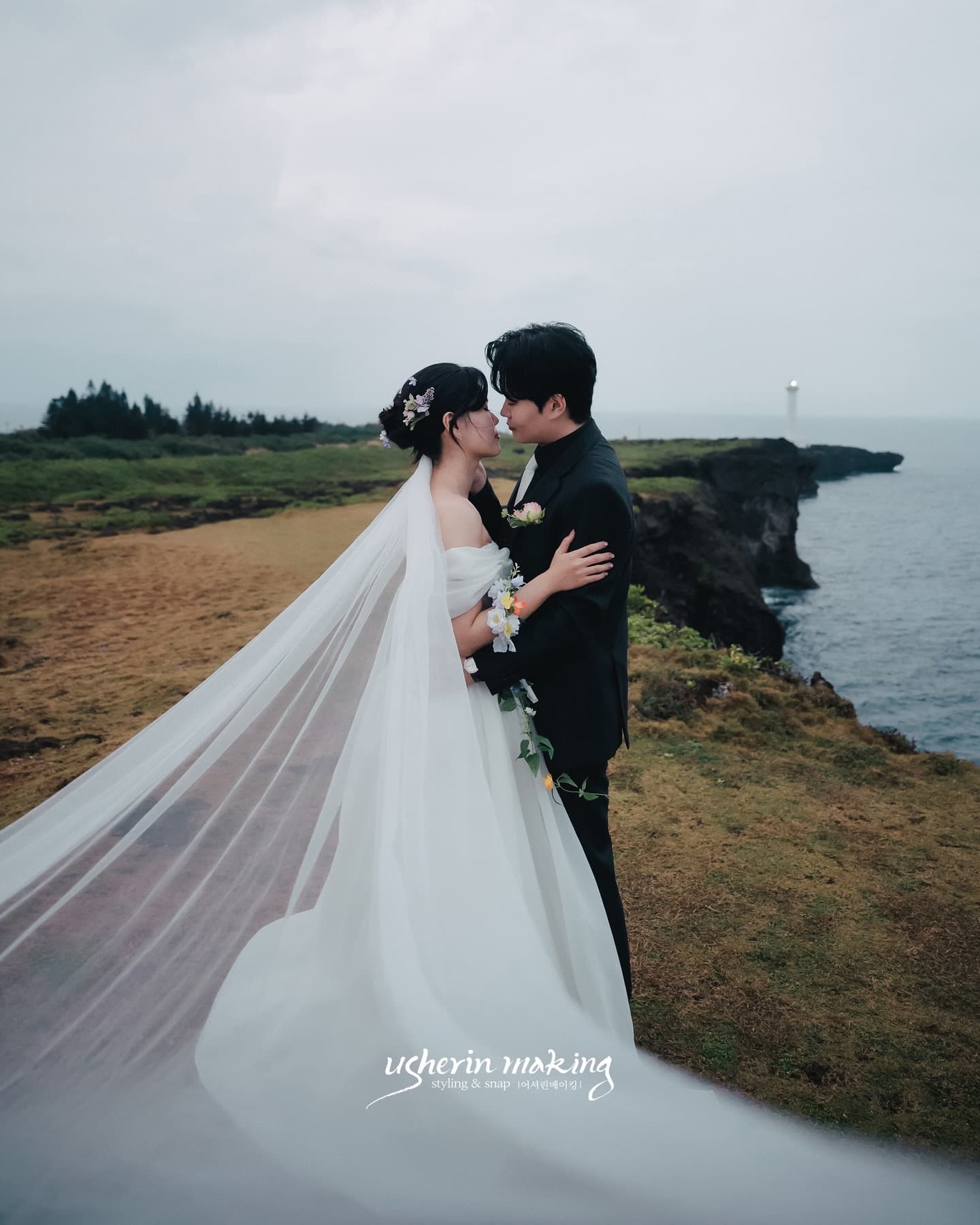 灯台を望む雨の岬でベールをなびかせ寄り添う新郎新婦、沖縄の前撮り