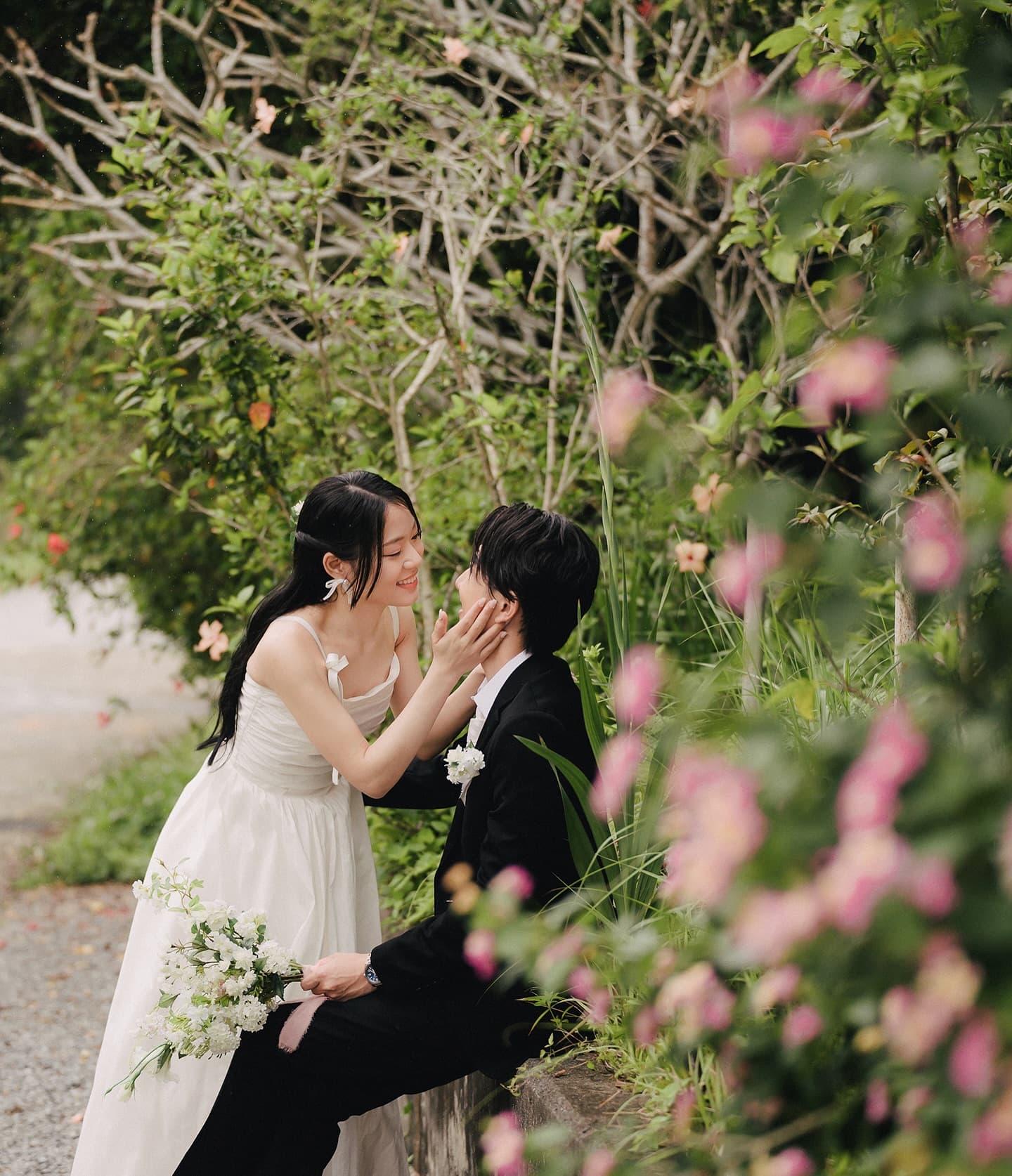 花咲く小道で新郎の頬に手を添えて微笑む新婦、緑あふれる沖縄