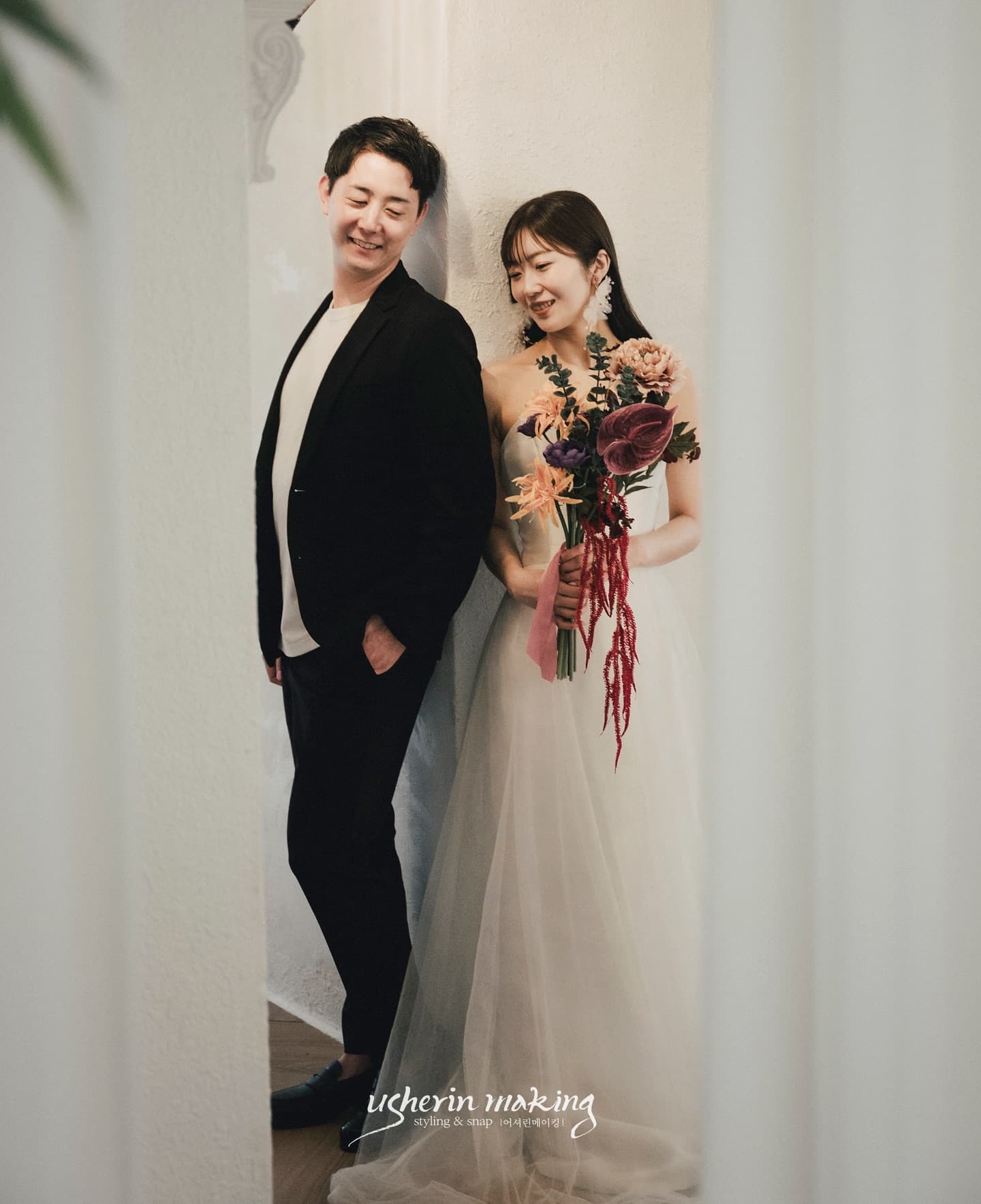 白い壁にもたれる新郎とブーケを持つ新婦、韓国風スタジオウェディングフォト