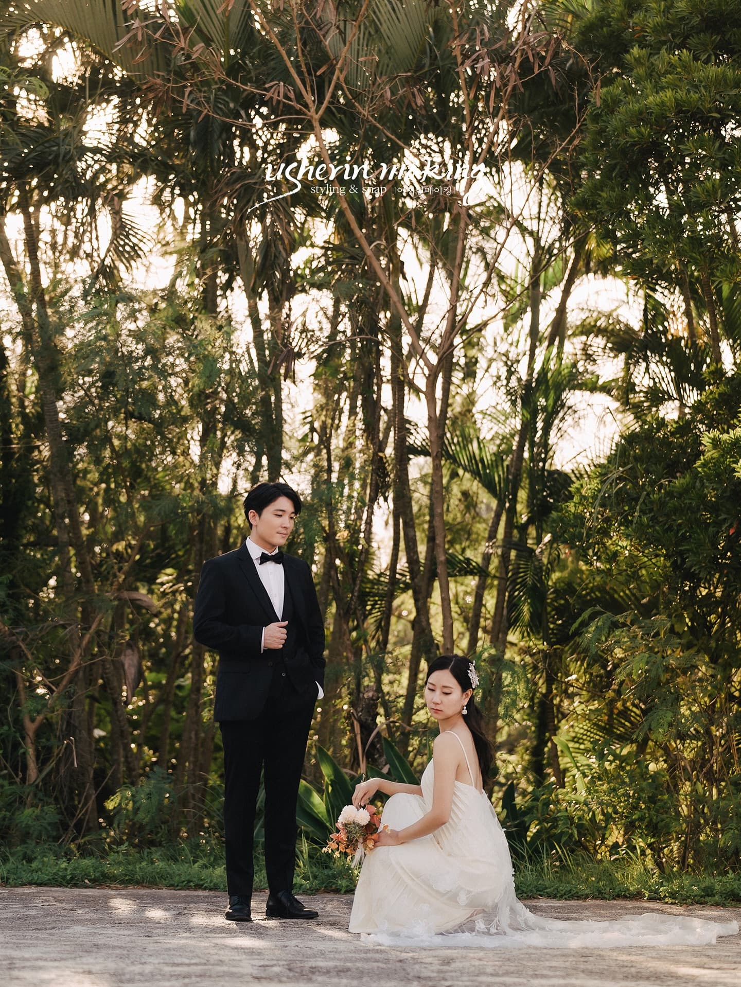 Bride with bouquet and tuxedoed groom under tall tropical trees in Okinawa