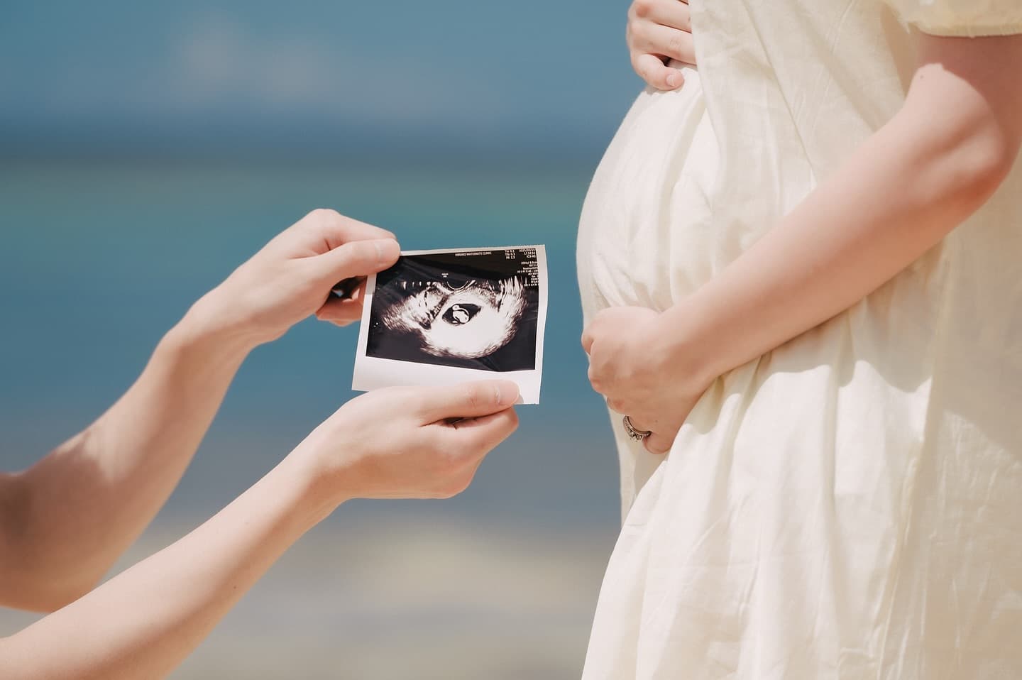 妊婦のお腹の前でエコー写真を掲げる手元、沖縄の海辺のマタニティフォト