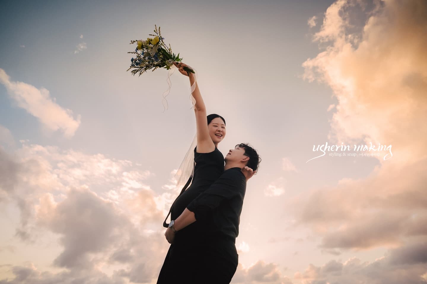 Bride in a black dress raising her bouquet high as the groom holds her under a sunset sky in Miyakojima