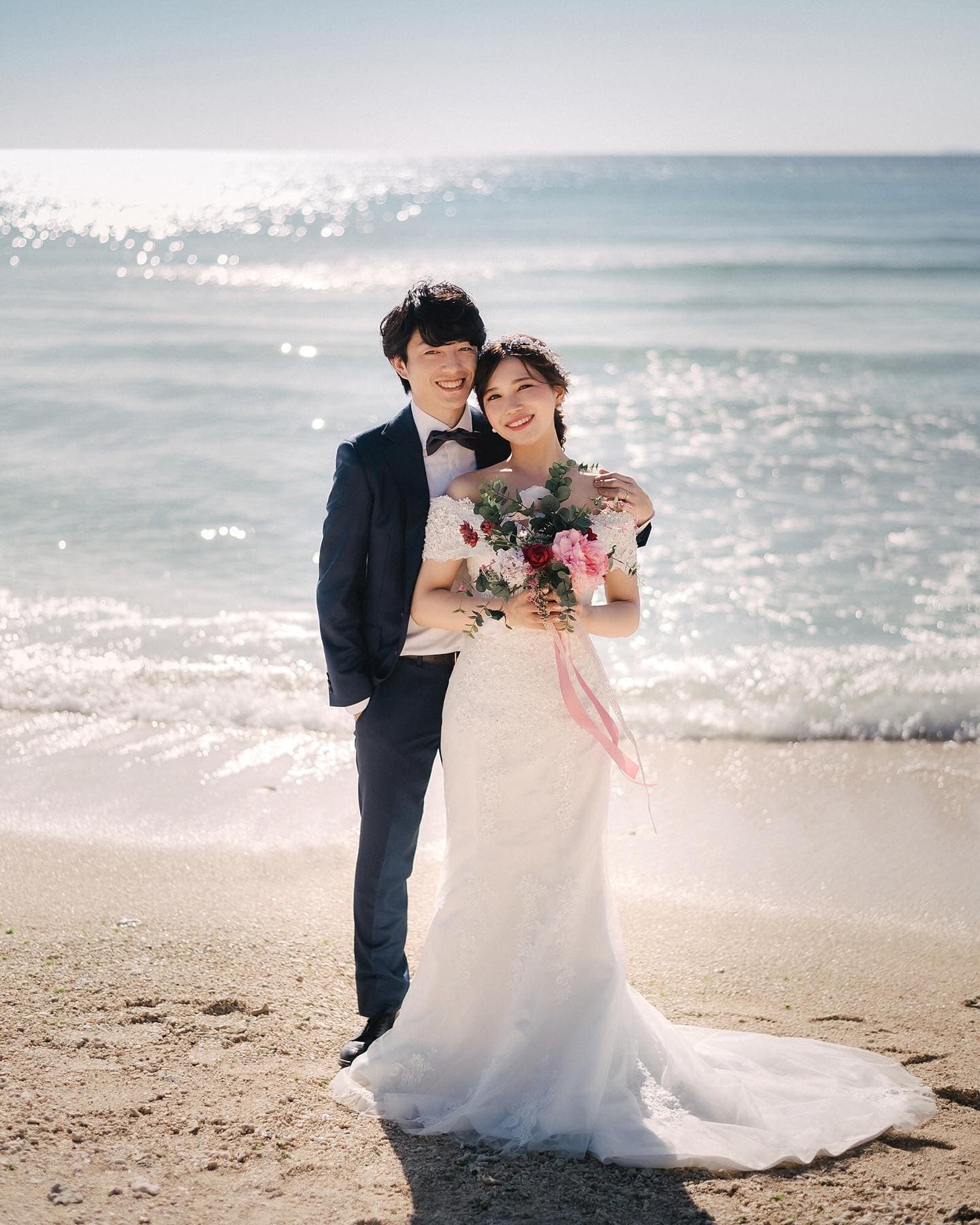 A smiling couple embrace on an Okinawa beach with the sparkling sea behind them