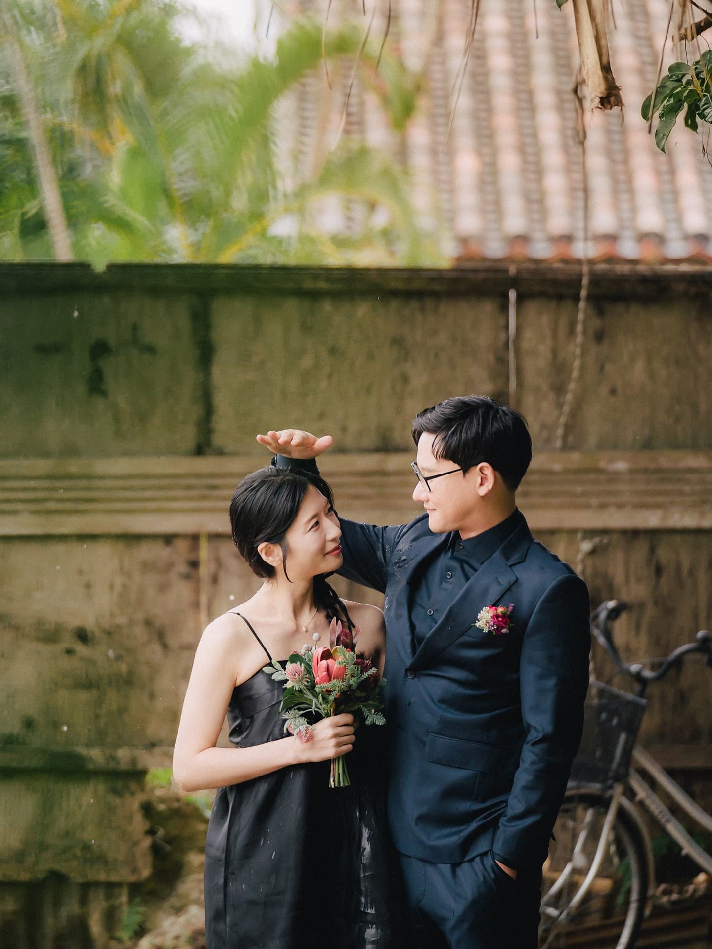 Groom shielding the bride from rain by an old red-tiled wall, Okinawa