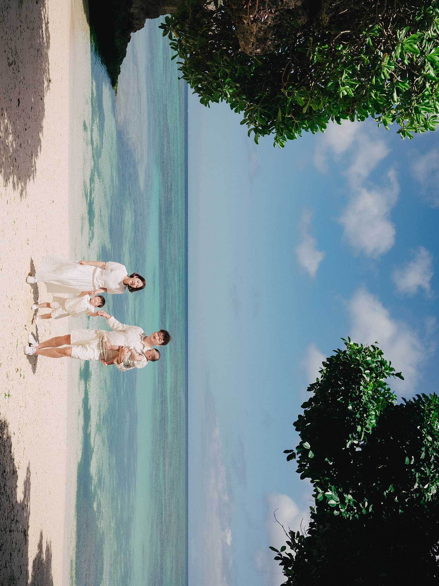 Family in white outfits holding hands on a white-sand Okinawa beach under a blue sky