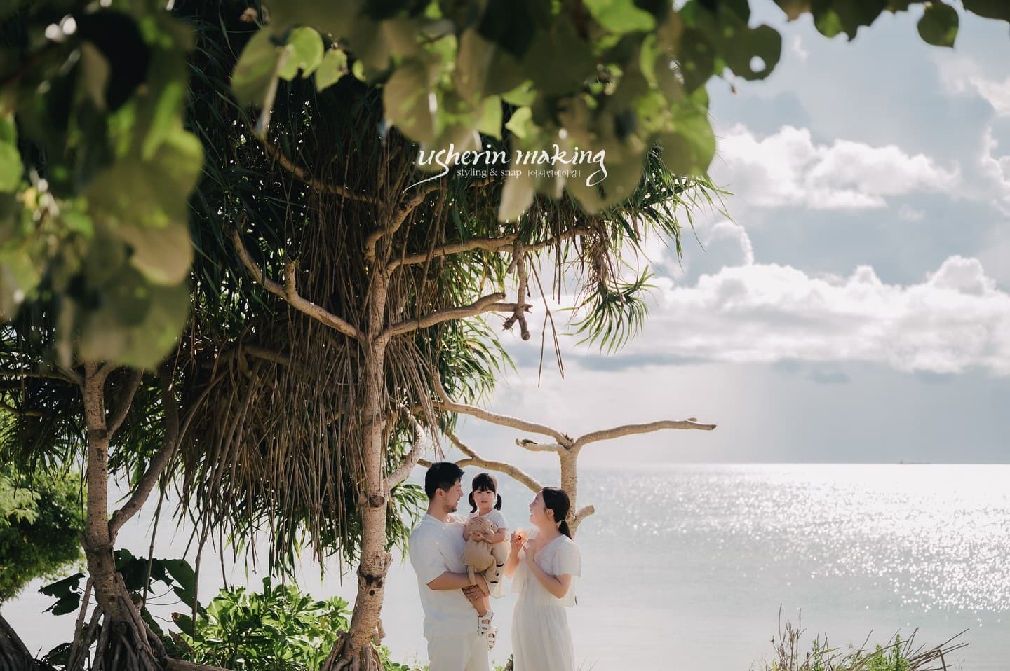 Family holding their baby under lush trees by the sparkling sea on a sunny Okinawa day