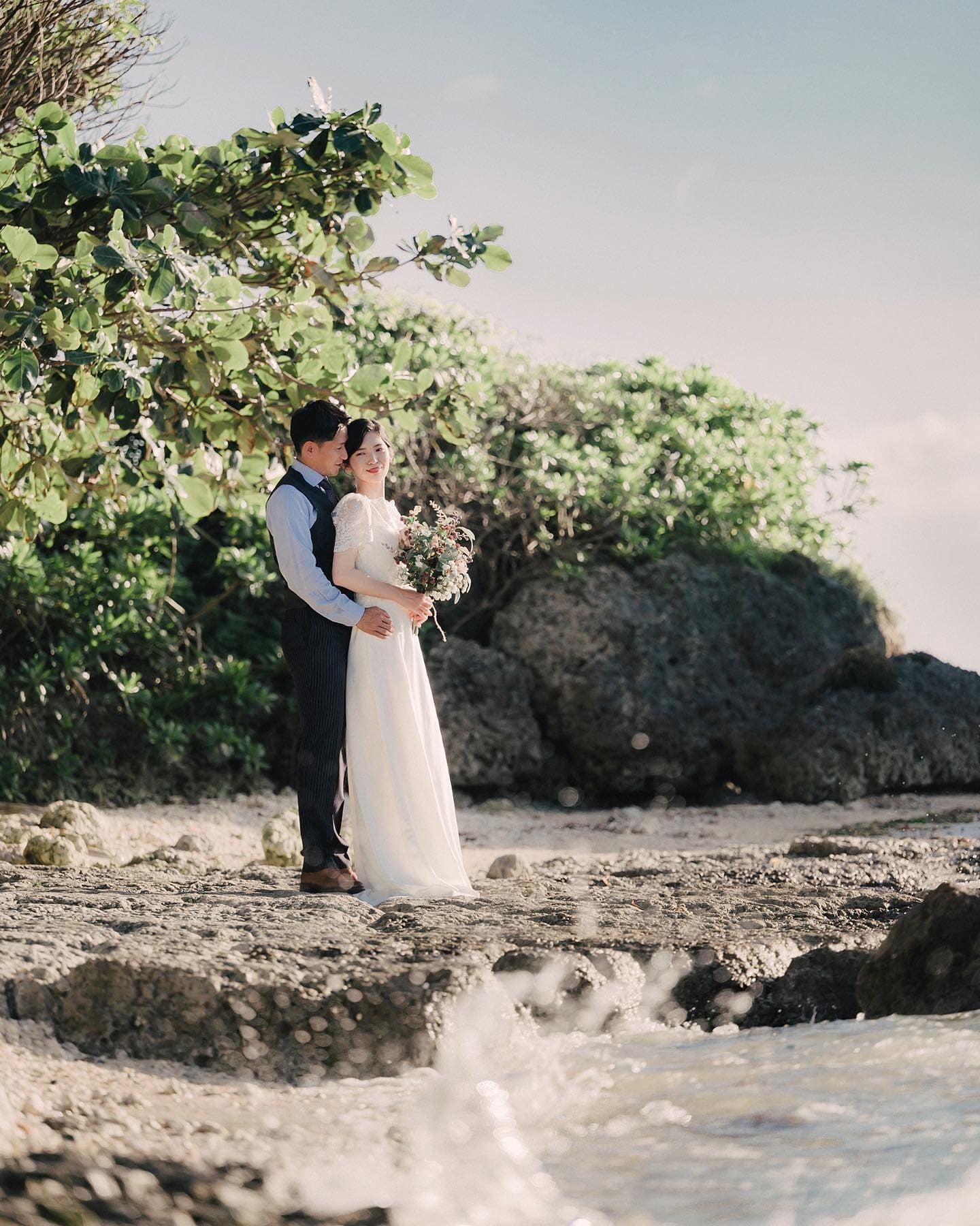 Groom embracing his bride from behind on a lush rocky Okinawa beach