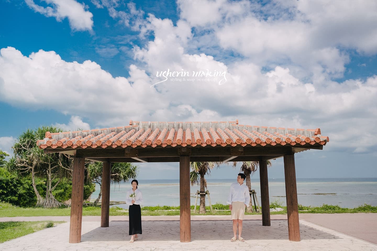 Couple standing beneath a red-tiled pavilion by the sea under a blue Okinawa sky