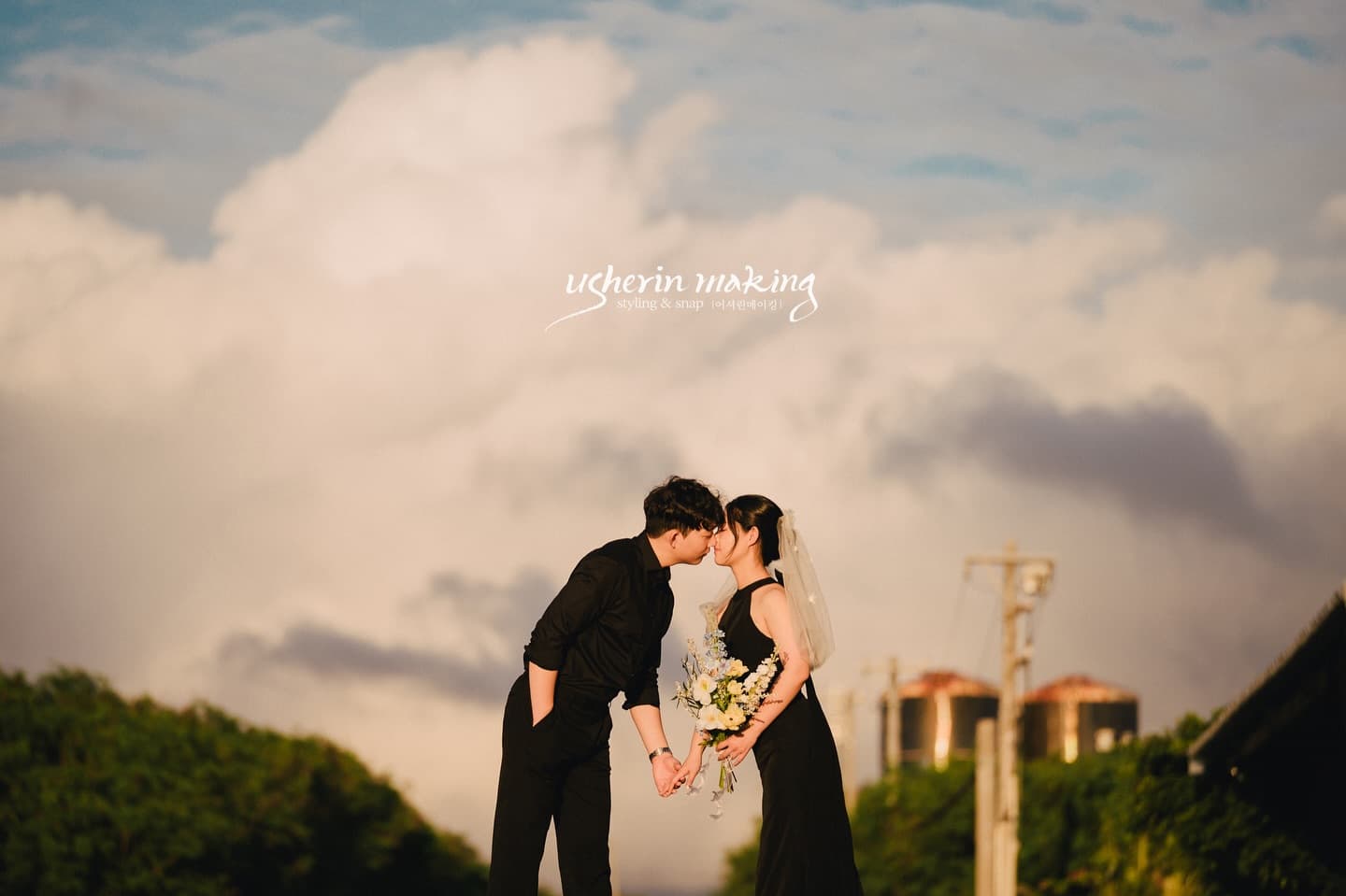 Couple dressed in black kissing on a greenery-lined road beneath towering clouds in Miyakojima