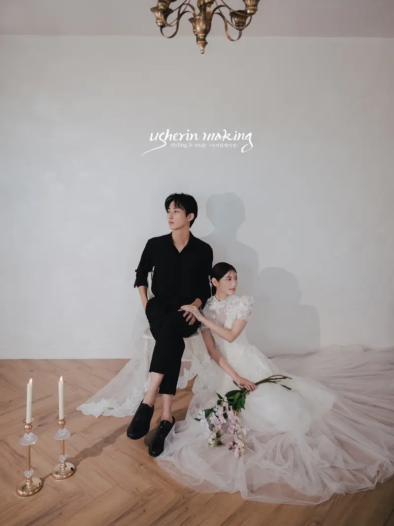 Groom in black seated beside the bride on the floor with a pink bouquet under a chandelier
