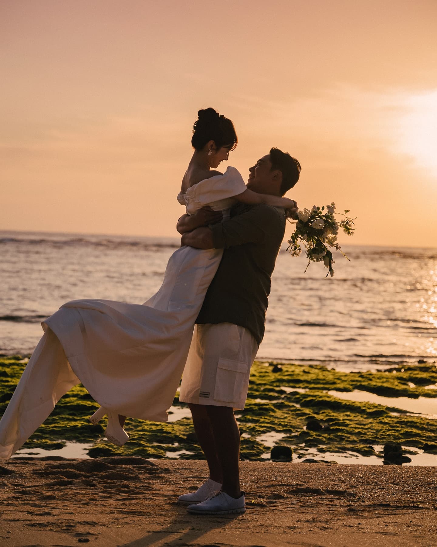 オレンジ色の空の下、抱き上げられてドレスが揺れる花嫁、沖縄の夕暮れ