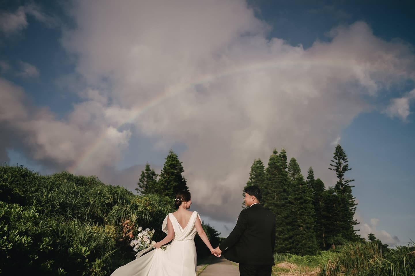 Bride and groom walking hand in hand toward a rainbow arching over the Okinawa sky