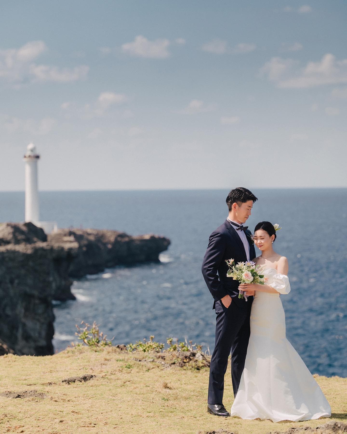 白い灯台と青い海を望む岬に立つ新郎新婦、沖縄