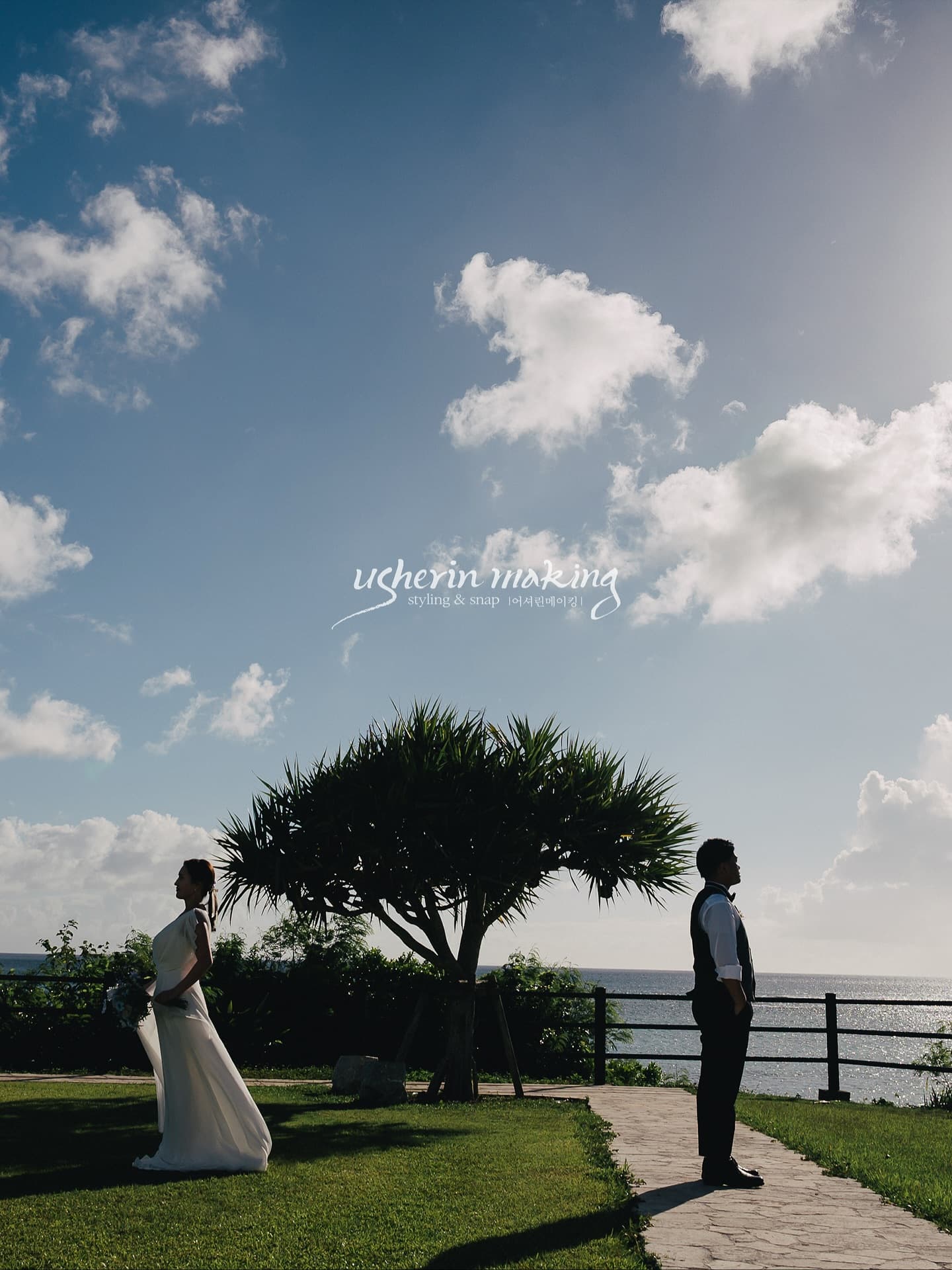 Bride and groom on a seaside lawn beneath a tall tree and wide blue Okinawa sky
