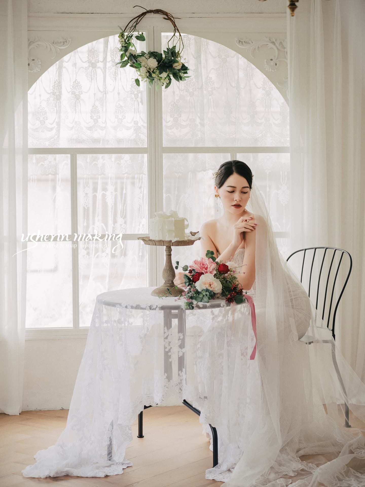 A veiled bride sits at a lace-draped table with cake by an arched window