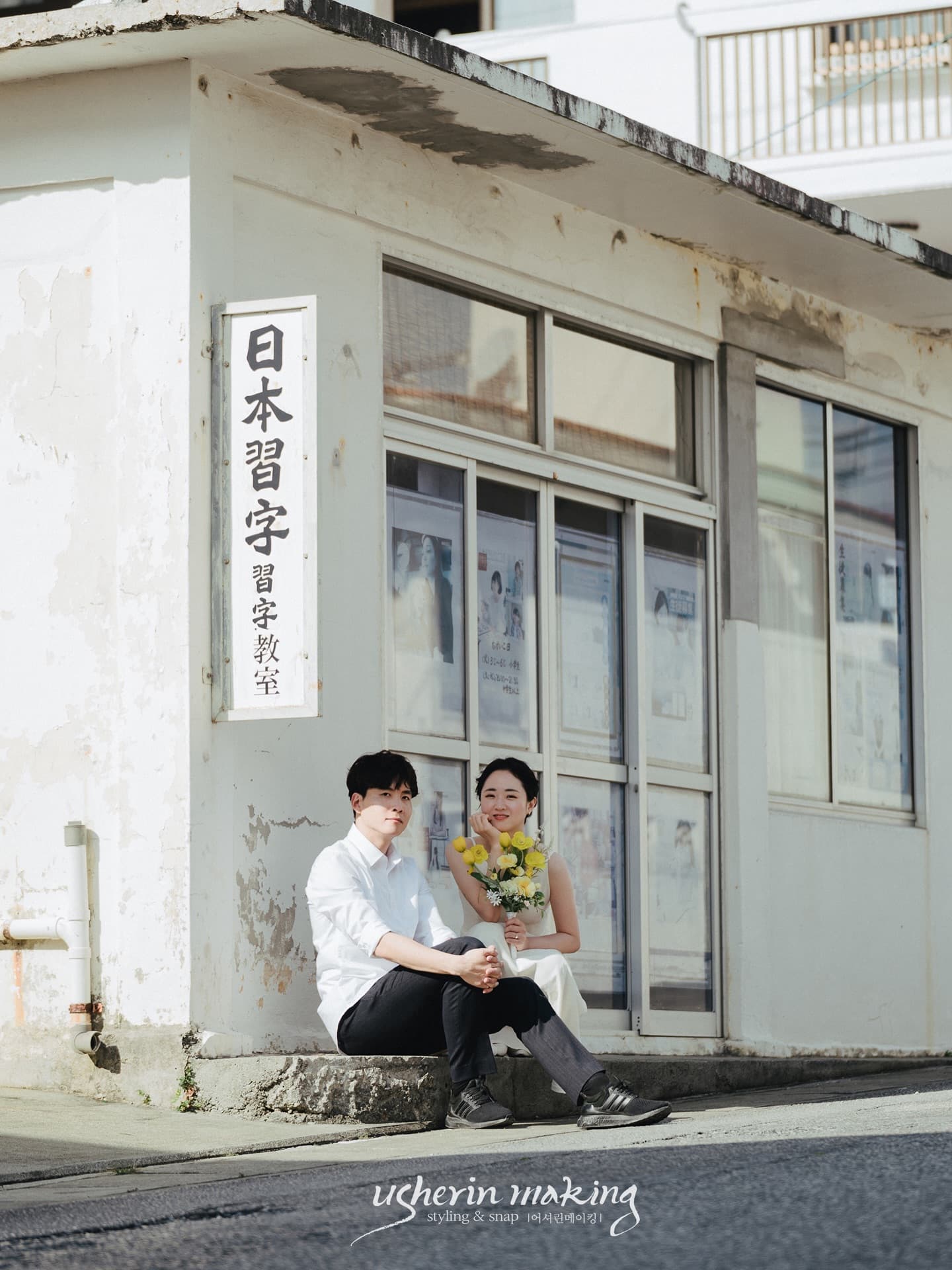 Couple sitting on the step of an old calligraphy school in an Okinawa town, holding yellow flowers