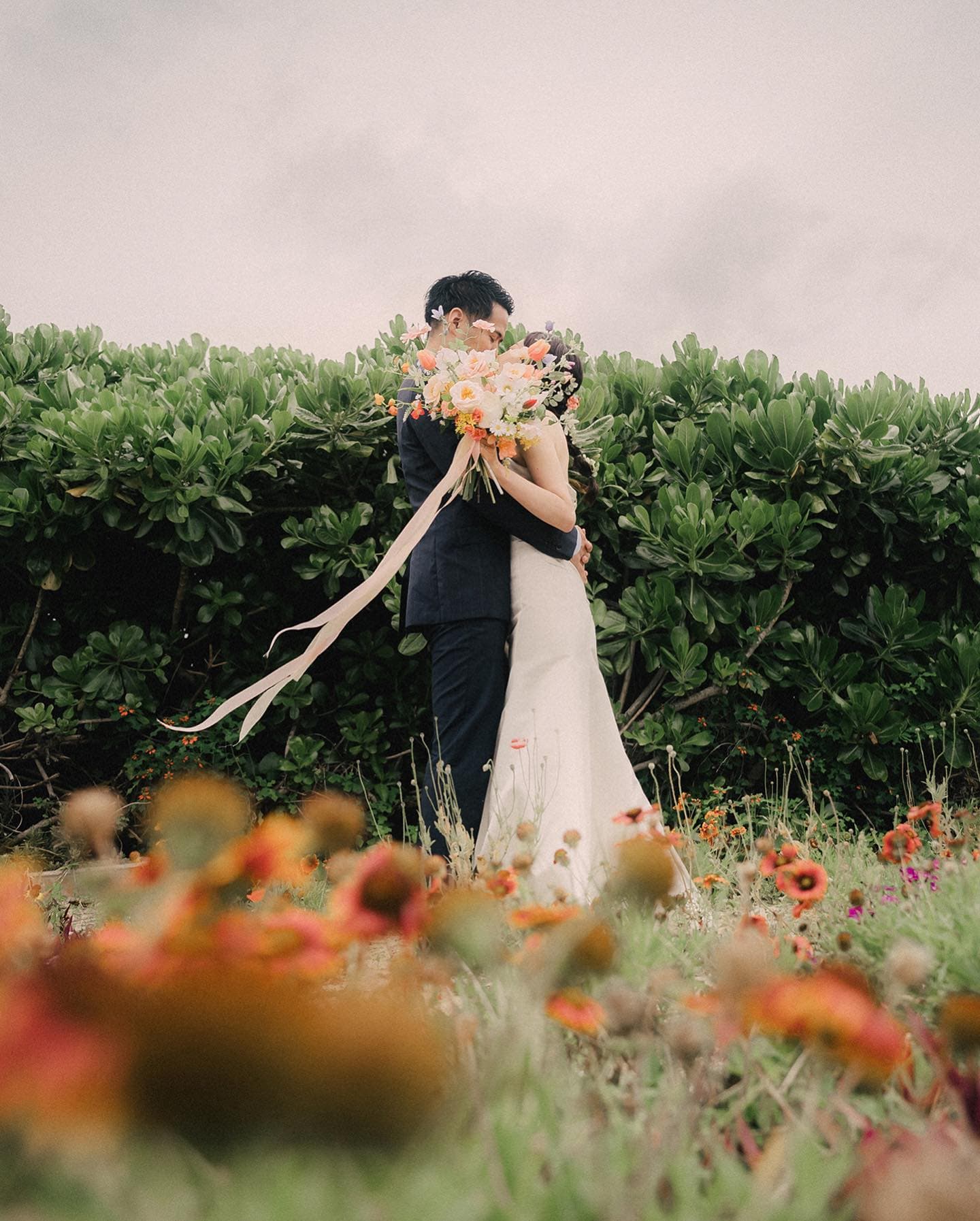 Bride and groom embracing with a large orange bouquet before a green hedge in Okinawa