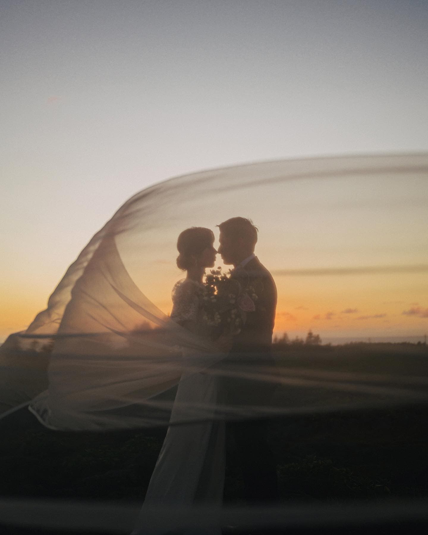 Couple facing each other inside a sweeping arc of veil against the dusk sky
