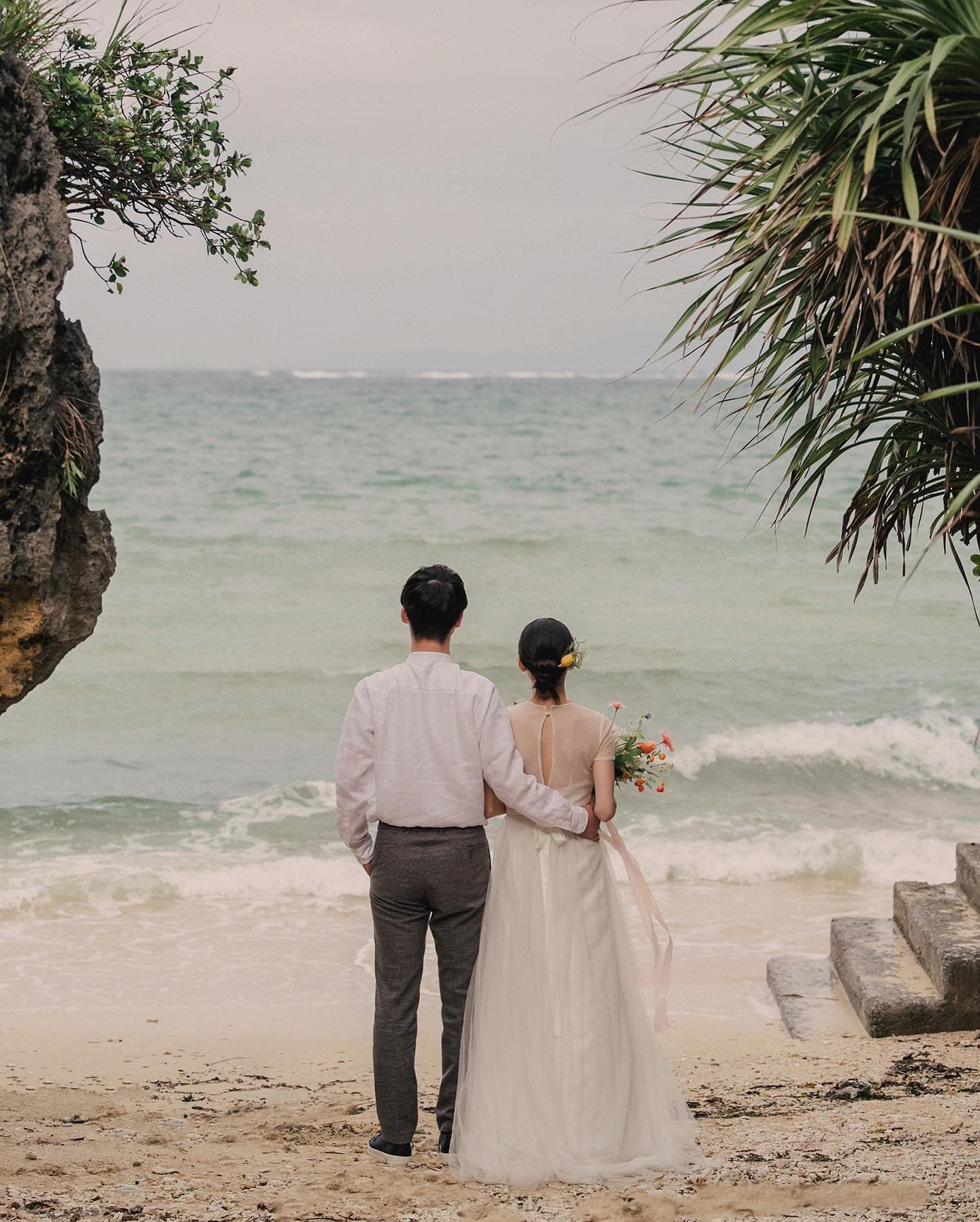 Bride and partner looking out to sea from a sandy beach on a cloudy Okinawa day