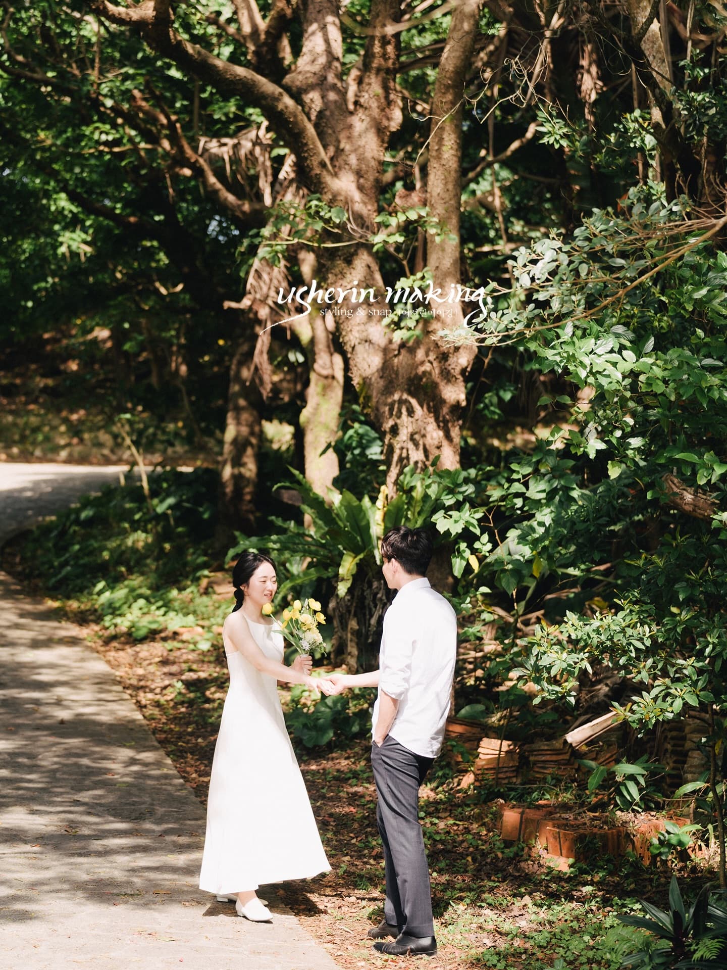 Couple facing each other on a shaded forest path in Okinawa, the bride holding yellow flowers