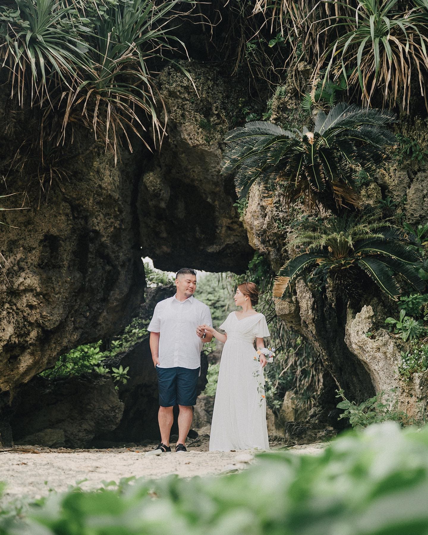 Couple in white standing beneath a rock arch framed by tropical plants in Okinawa