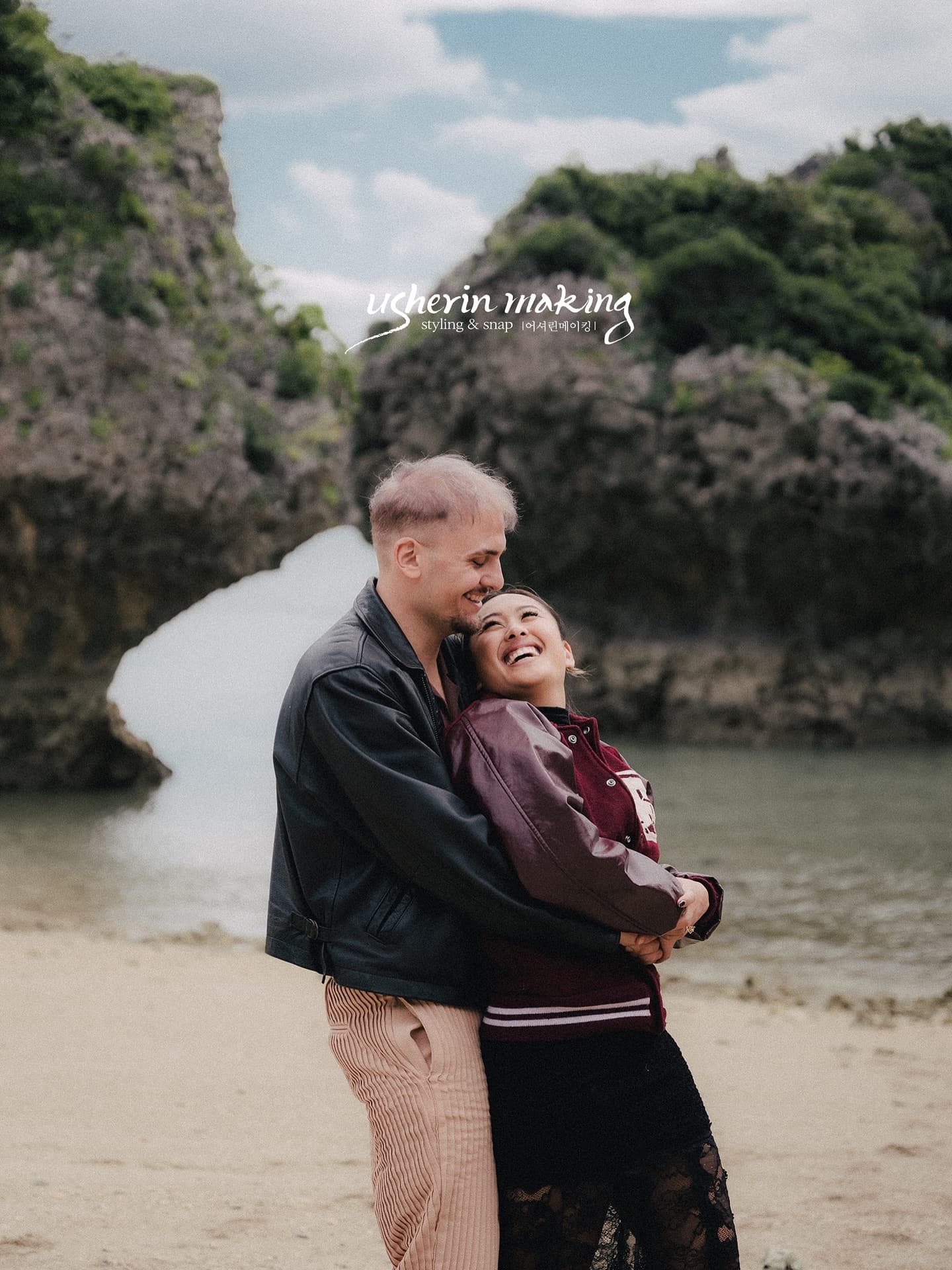 Couple in leather jackets embracing and laughing at a rocky Okinawa cove on a cloudy day