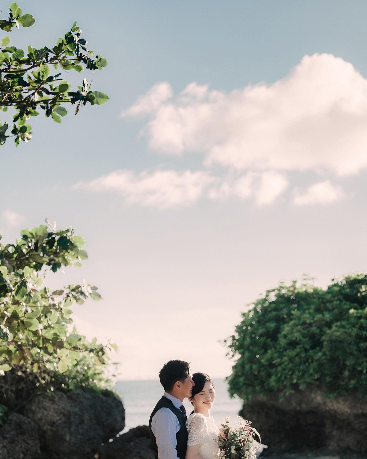 Bride and groom framed by green rocks under a wide sky on the Okinawa coast