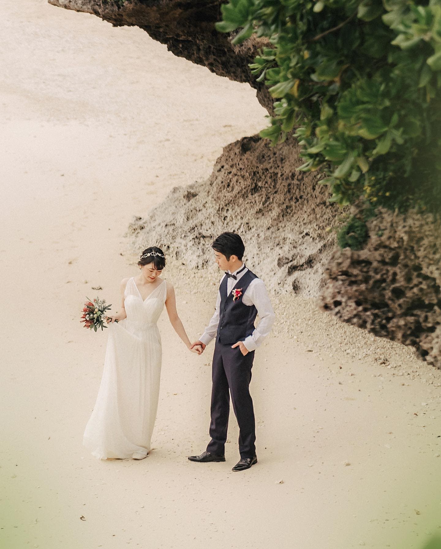 Bride and groom strolling hand in hand across pale sand past a mossy rock in Okinawa