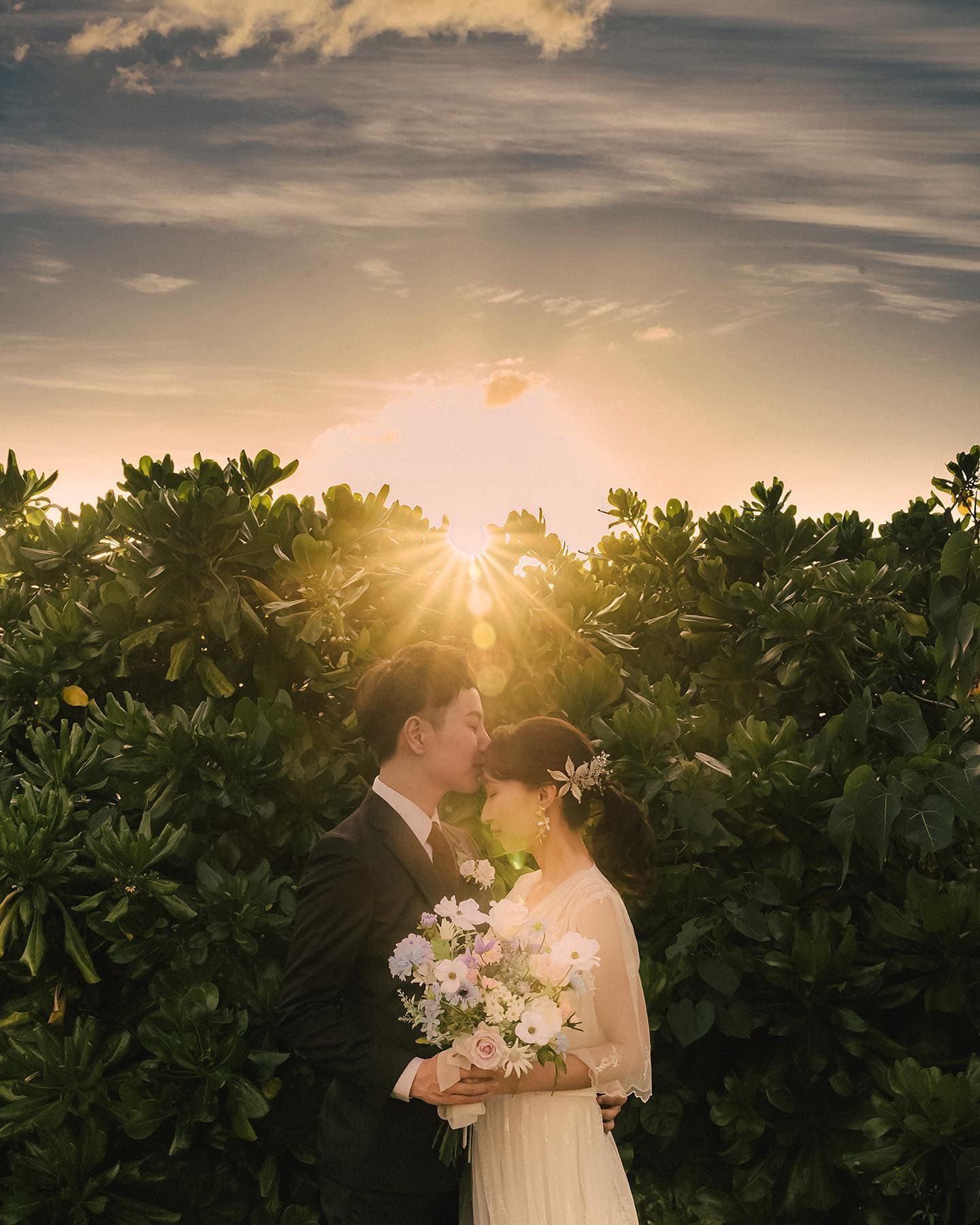 夕日が輝く緑の茂みの前で花嫁の額にキスする新郎、沖縄の前撮り