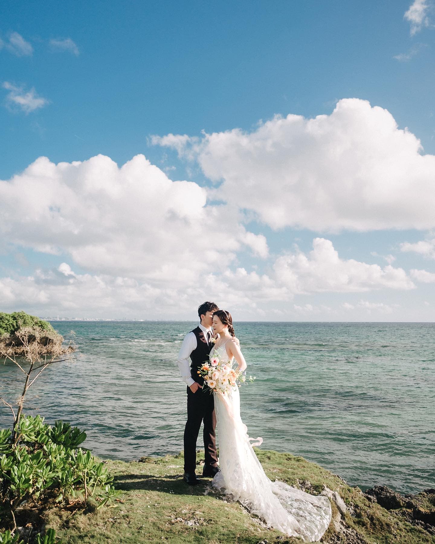 Bride and groom embracing on a green cape above the emerald sea in Okinawa