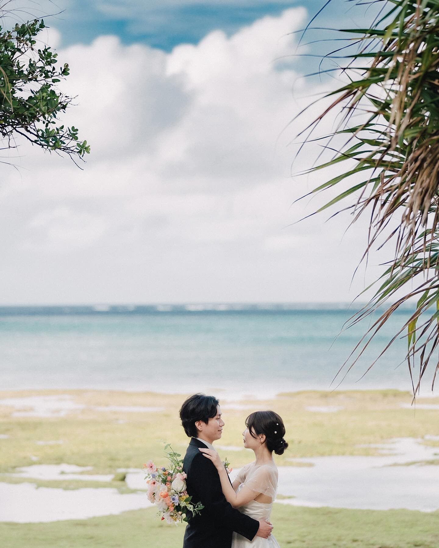 海と青空を背に向かい合って抱き合う新郎新婦、ヤシの葉が縁取る沖縄
