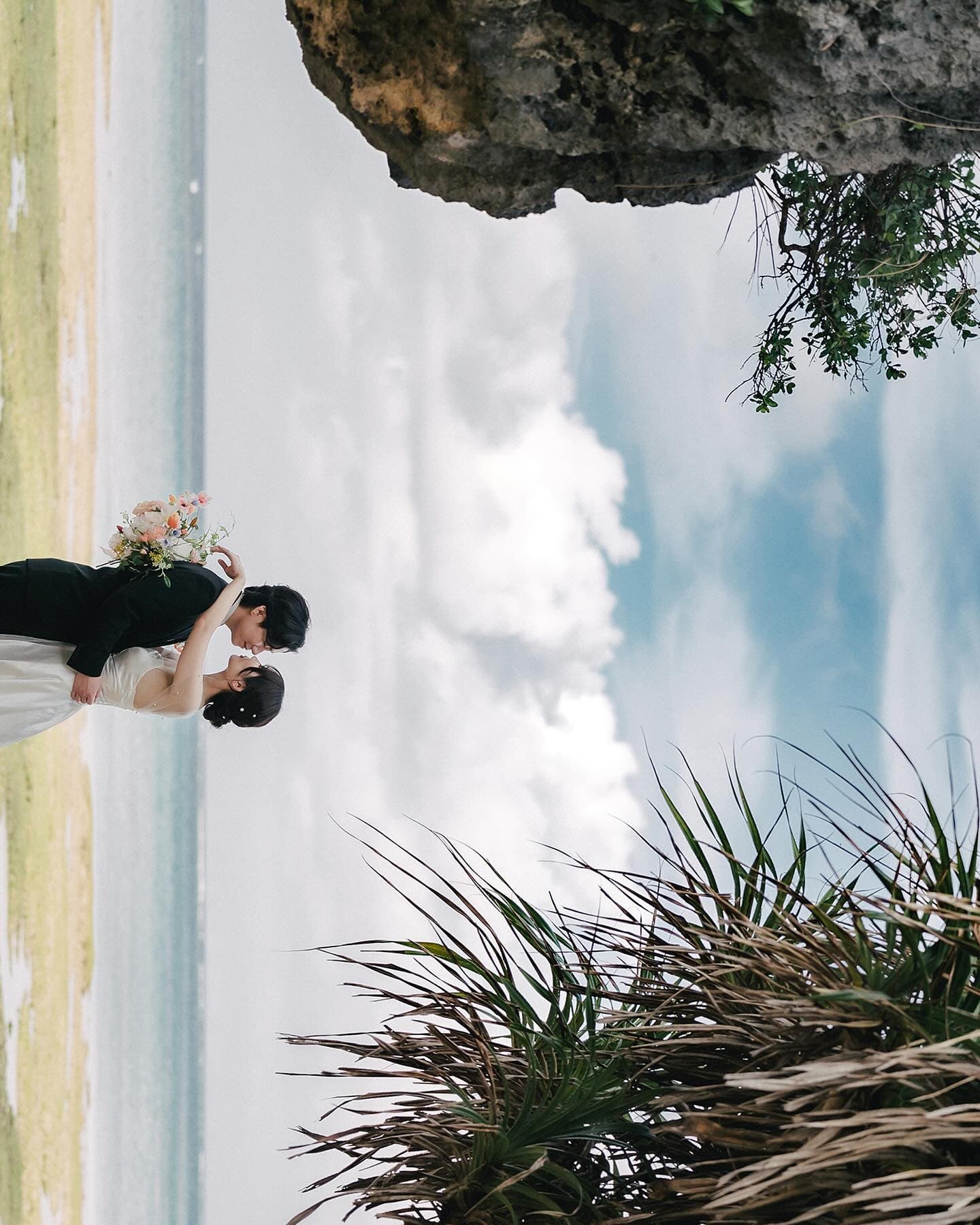 岩壁の下で花束を掲げてキスする二人、雲の広がる沖縄の海辺