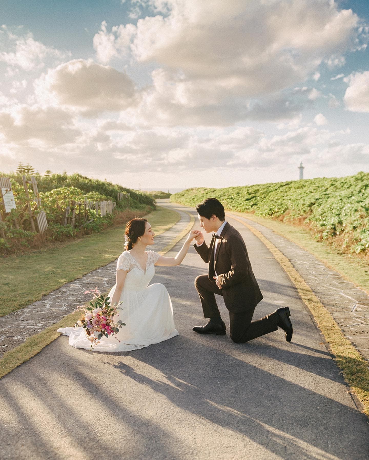 Groom kneeling before his bride on a greenery-lined path under a cloud-dotted sky