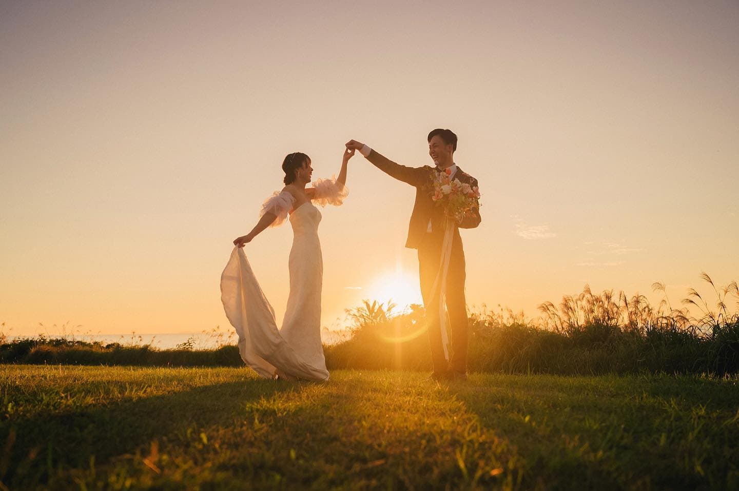 Bride and groom dancing hand in hand on a grassy field at sunset in Okinawa