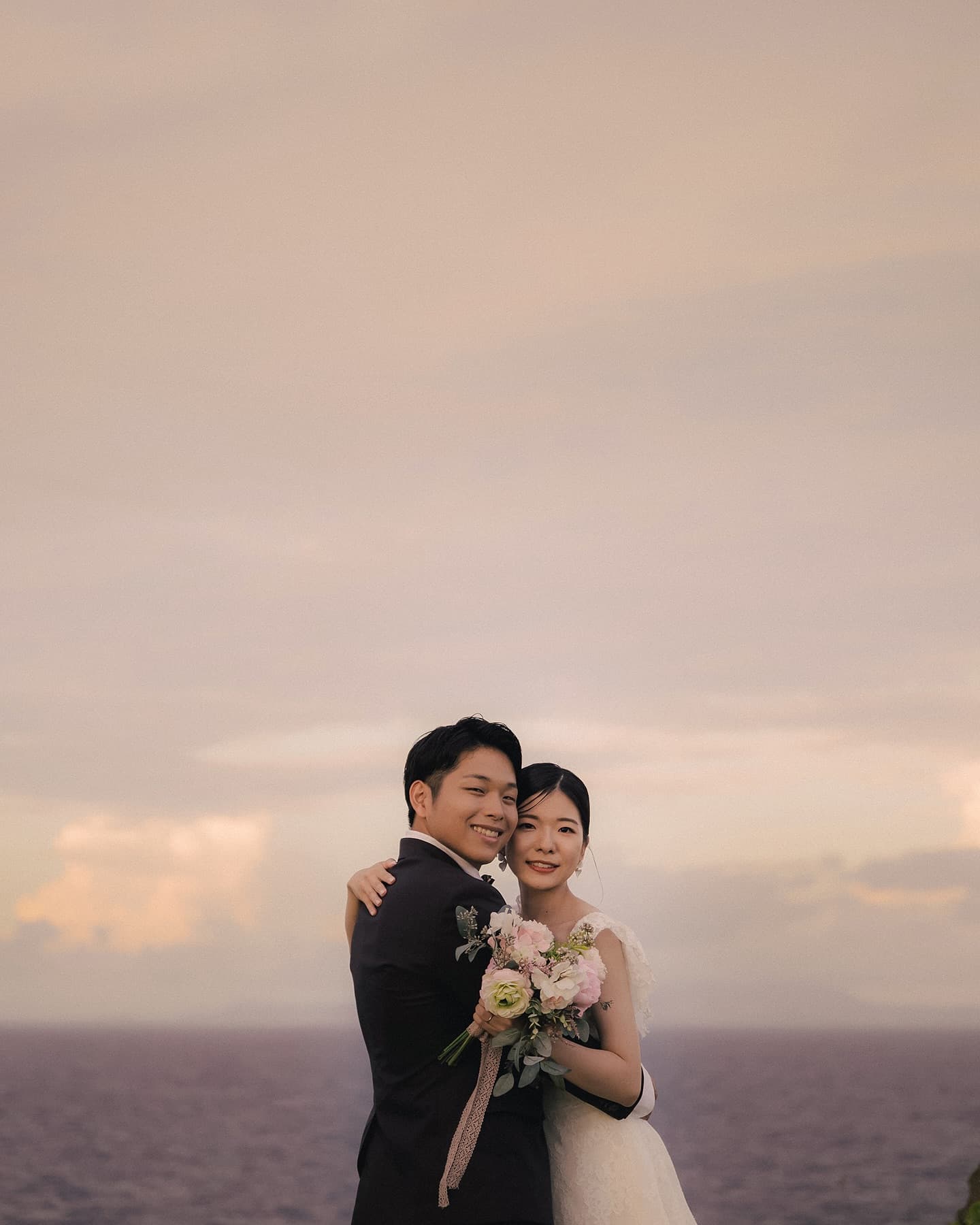 ピンク色に染まる夕空の海辺でブーケを手に抱き合う新郎新婦、沖縄