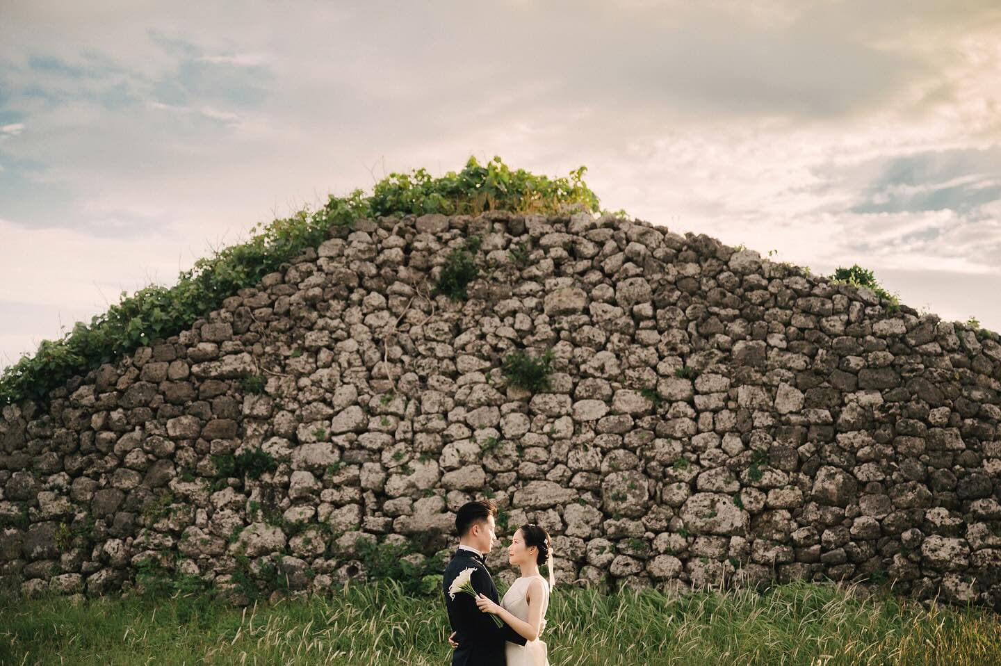 A wedding couple embraces before old stone ruins under a cloudy Okinawa sky
