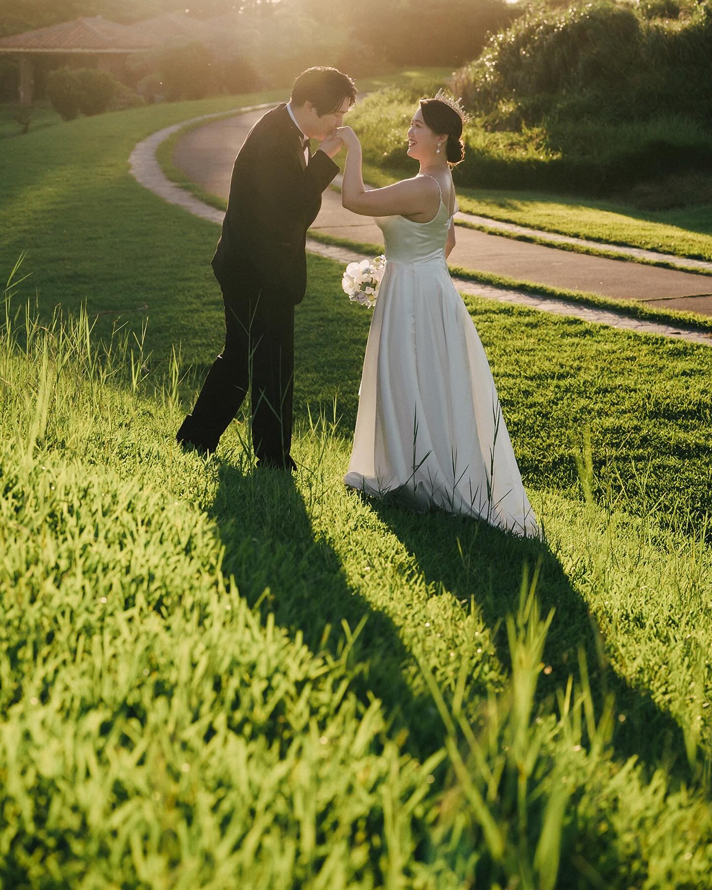 Groom kissing the bride's hand on a sunlit green lawn in Okinawa