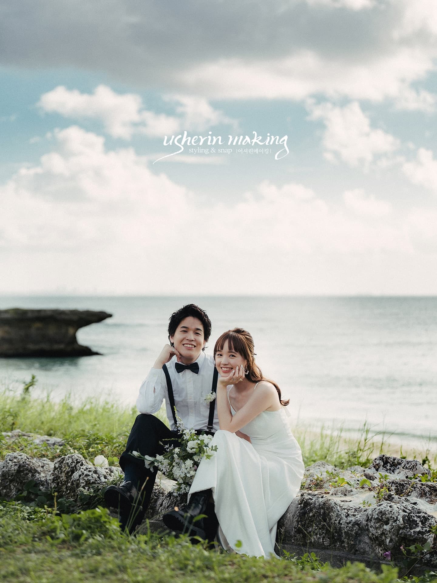 海辺の岩に腰かけて微笑む新郎新婦、白い雲広がる沖縄の空