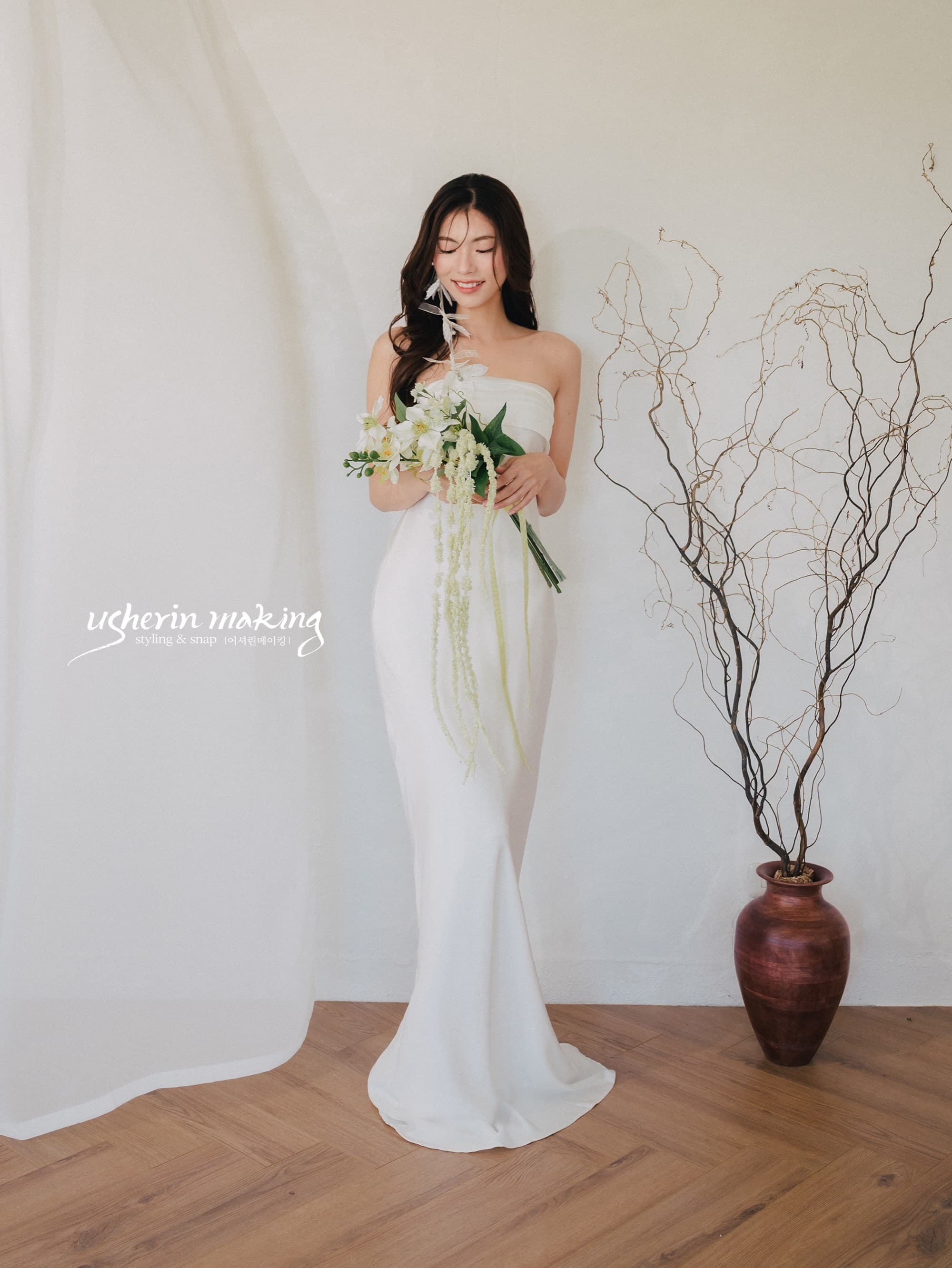 Bride in a slim white dress admiring her bouquet in a bright studio
