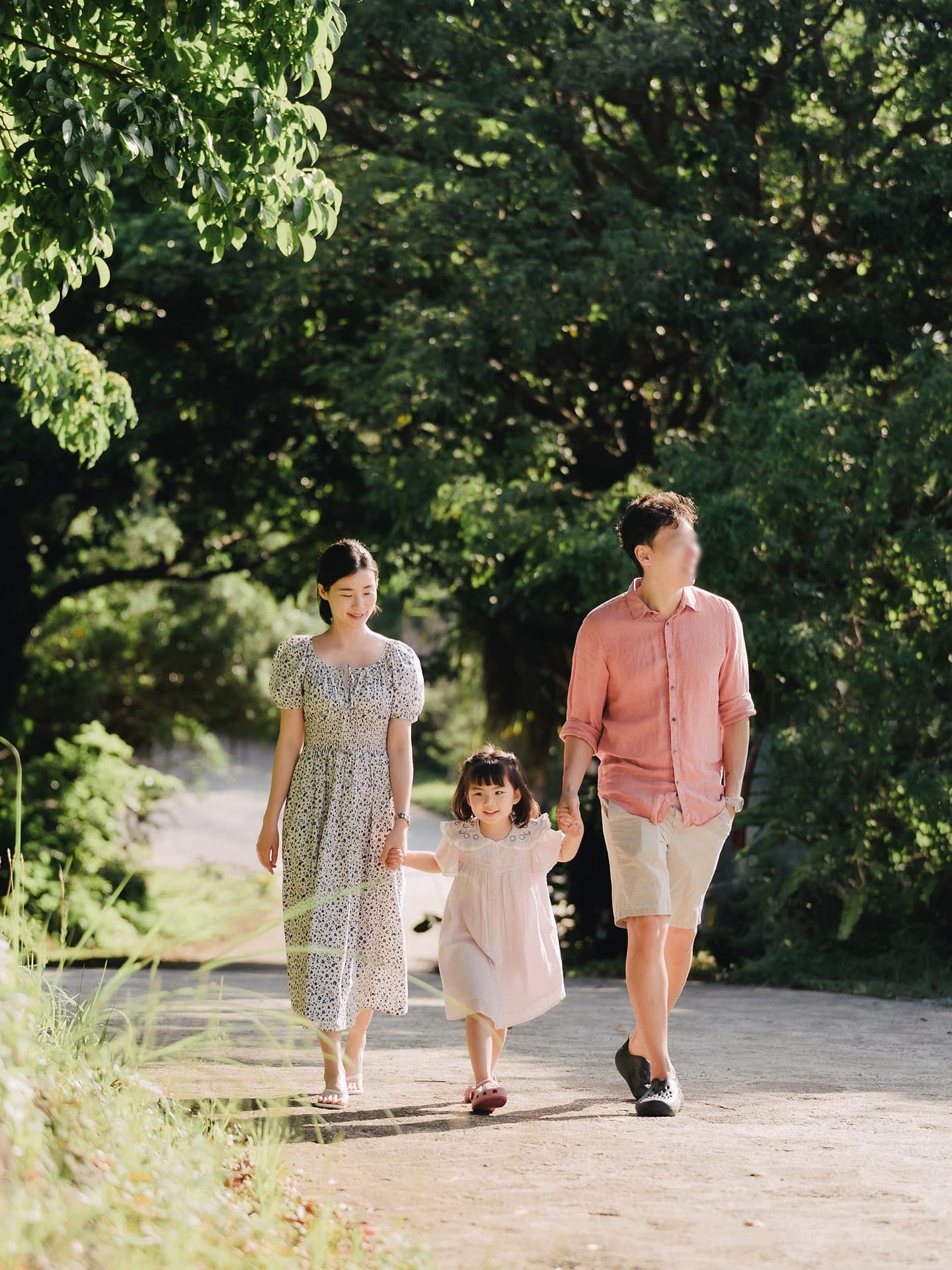 Family of three walking down a sunlit green lane holding their daughter's hands in Okinawa