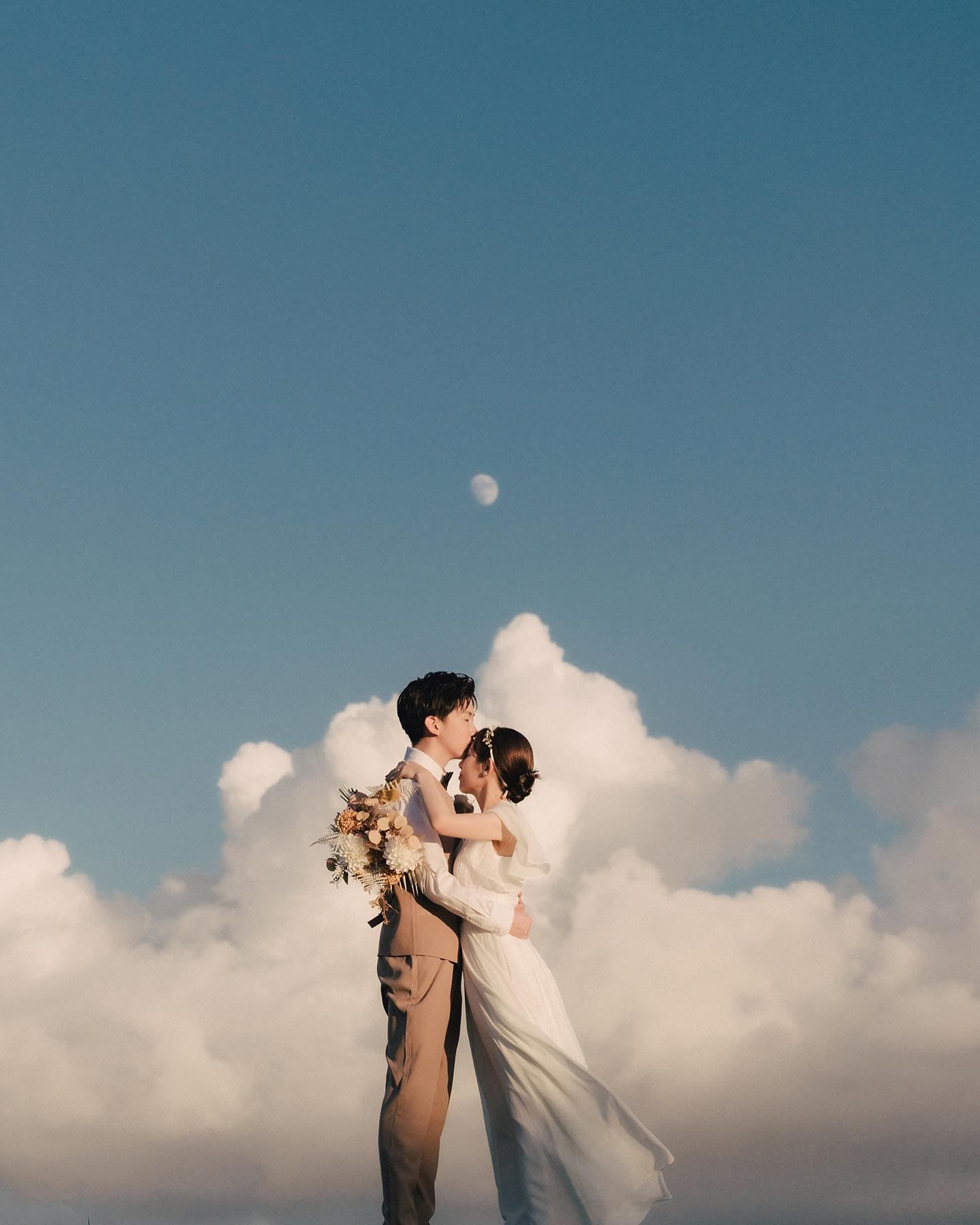 Couple embracing beneath a small moon and billowing white clouds in Okinawa