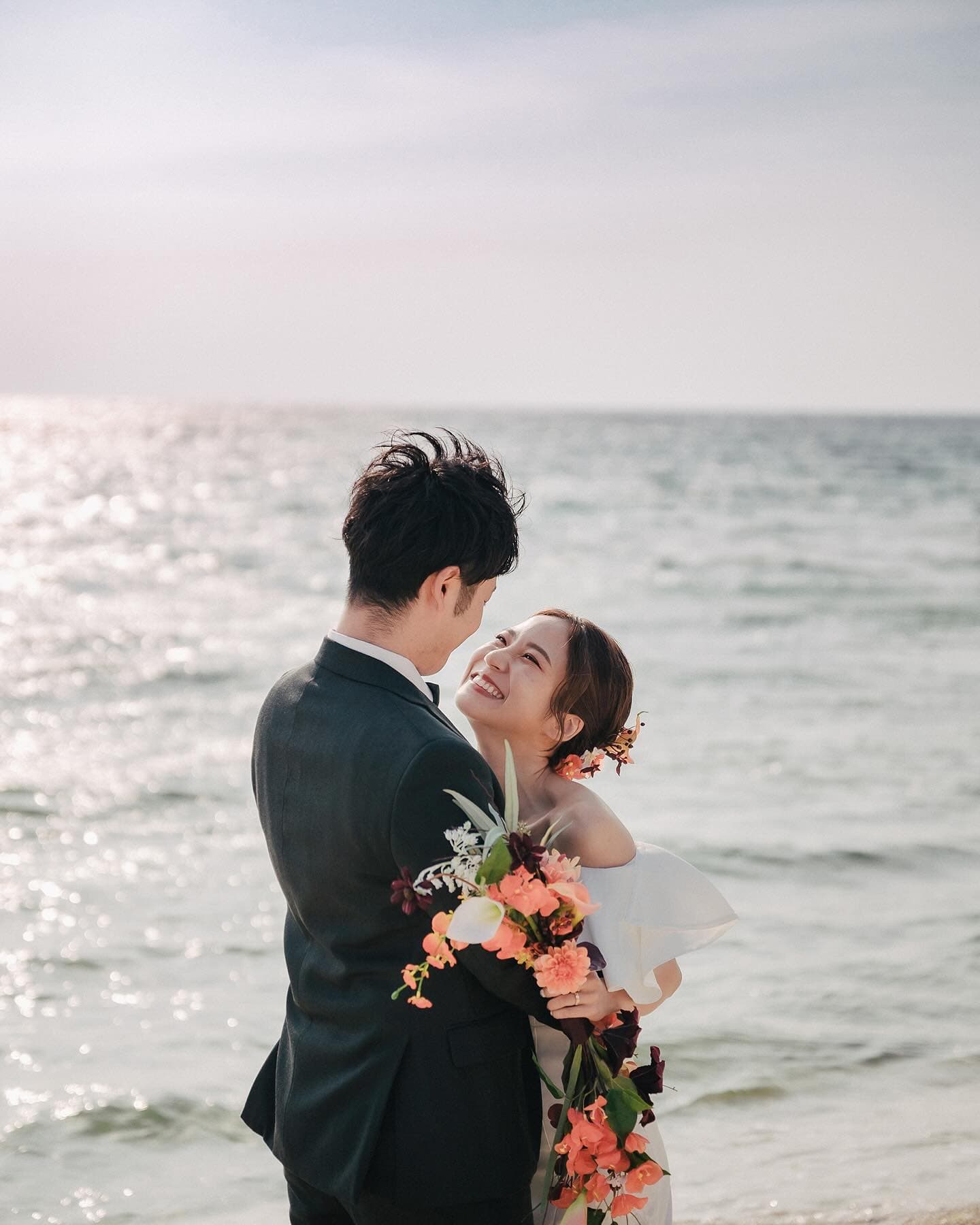 Bride laughing in the groom's arms with orange flowers by the sparkling Okinawa sea
