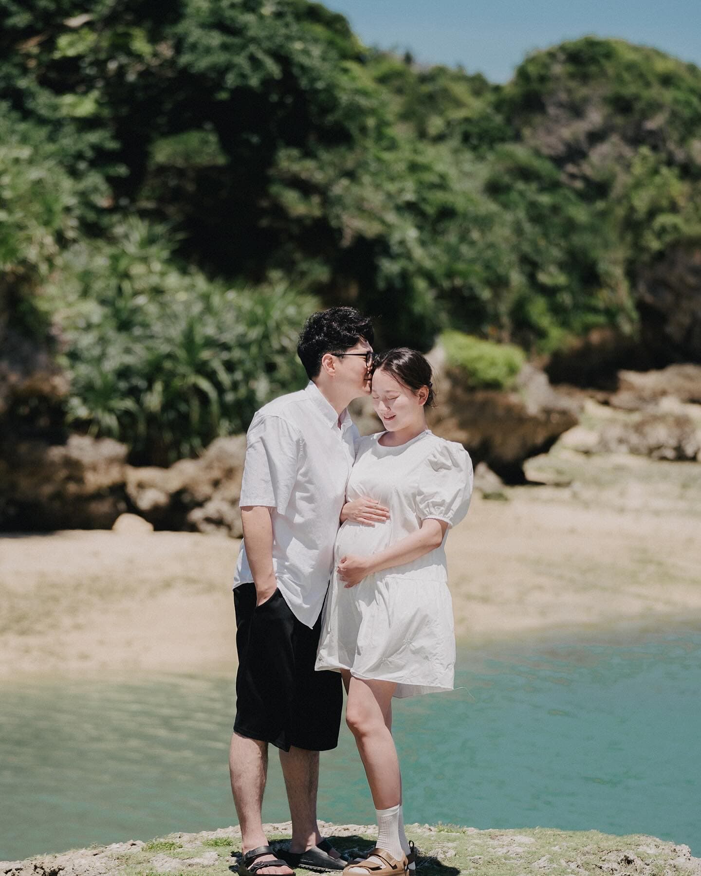 Husband kissing his pregnant wife's forehead by an emerald cove in Okinawa